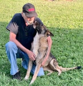 A man with a black baseball cap, black t shirt, and blue jeans, is kneeling beside a rare Panda German Shepherd which has a white chest and white paws with a black mask. The German Shepherd seems to be whispering in the mans ear as they relax on a sunny day in the green grass