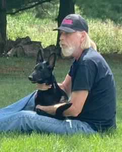 A man sitting in the grass, legs out in front of him. He is holding an AKC Black Bear German Shepherd®