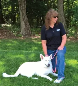 A woman with long brown hair, wearing blue jean, a dark blue polo shirt with white letters across the chest, and white sneakers is kneeling beside a white German Shepherd who is lay down in the green grass. The woman is wearing sunglasses as the sun is shining brightly on the pair. Trees are in the background.