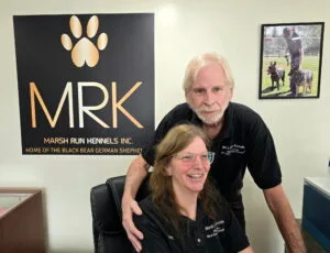 Image of Betsy Stevens and Bart Stevens. Owners of Marsh Run Kennels Inc. Betsy is sitting at the reception area while Bart is leaning over her. Both are smiling and greeting a client. 