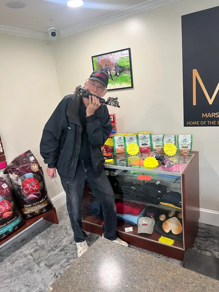 Dogman posing with a rope toy beside Marsh Run Kennels merchandise display featuring treats, apparel, and pet supplies.
